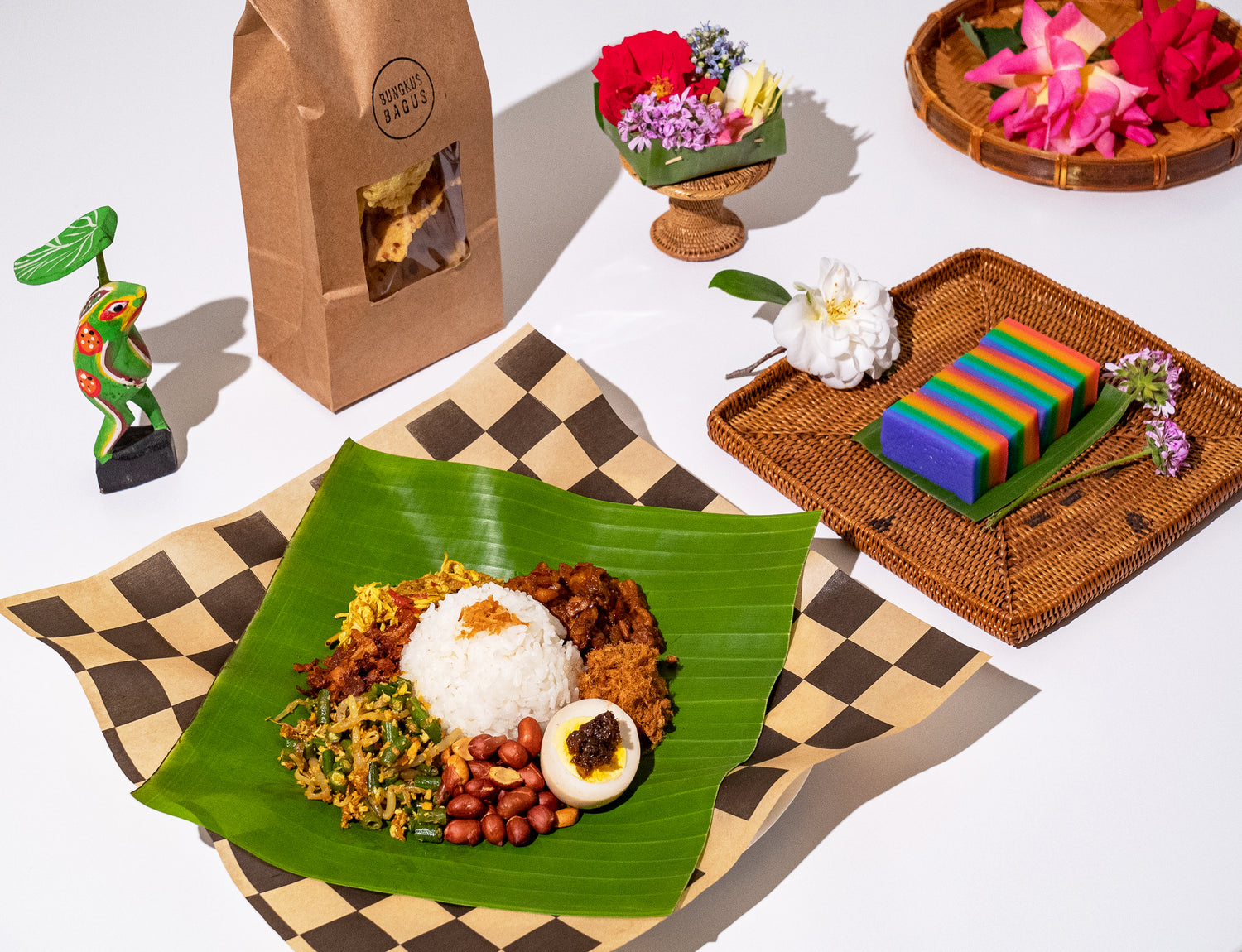 A Nasi Bungkus rests on a green banana leaf, along with Kueh Lapis dessert, Krupuk chips, and then tropical flowers.